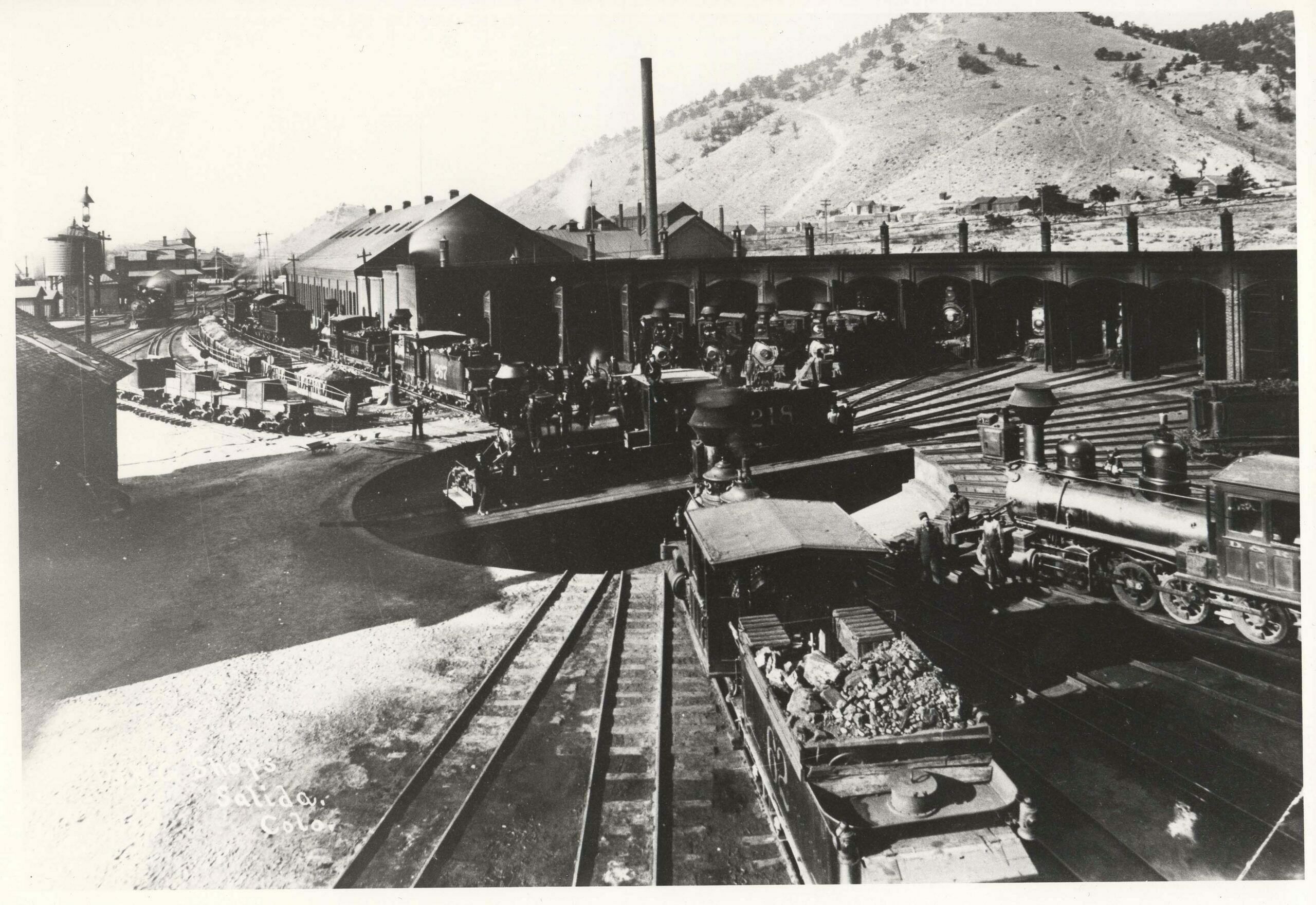 All 27 roundhouse tracks and nearly half of the Salida roundhouse itself, are visible in this photograph, taken sometime between 1890 and 1892. Nearly half of the stalls carry four rails to handle both gauges. The 62-foot turntable was used until 1909, when it was replaced with an 80-foot model. Moving counter-clockwise from the roundhouse lead nearest the photographer, the following locomotives are visible: •    narrow-gauge Engine 62, a Baldwin Class 56 2-8-0 (notice the irregular size of coal in its tender) •    narrow-gauge Grant Class 60 (C-16) 2-8-0, No.213 •    a narrow-gauge tender from a Class 60 locomotive •    an unidentifiable narrow-gauge locomotive •    two unidentifiable standard-gauge engines •    an unidentifiable narrow-gauge 2-8-0 •    narrow-gauge locomotive No. 404 •    an 1881 Baldwin Class 70 (C-19) 2-8-0 •    narrow-gauge No. 274, an 1882 Baldwin Class 60 (C-16) 2-8-0 •    No. 401, another Class 70 (C-19) 2-8-0 •    and two barely visible locomotives in the darkness of the roundhouse. Class 60 Grant 2-8-0 No. 218 rode on the turntable. Class 60 No. 267 and Class 70 No. 409, along with two unidentifiable standard-gauge engines, rested on the ashpit lead. Six gondolas full of ashes, along with one empty, were spotted next to the ashpit. Three drag flangers were next to the ashpit. Notice the water column next to engine No. 267. At left – looking into the distance – you can see the water tank, the Hotel Monte Christo, and the Salida depot. Notice the attractive arch doorways of each roundhouse stall. Tenderfoot Hill looms behind the roundhouse.  (2)