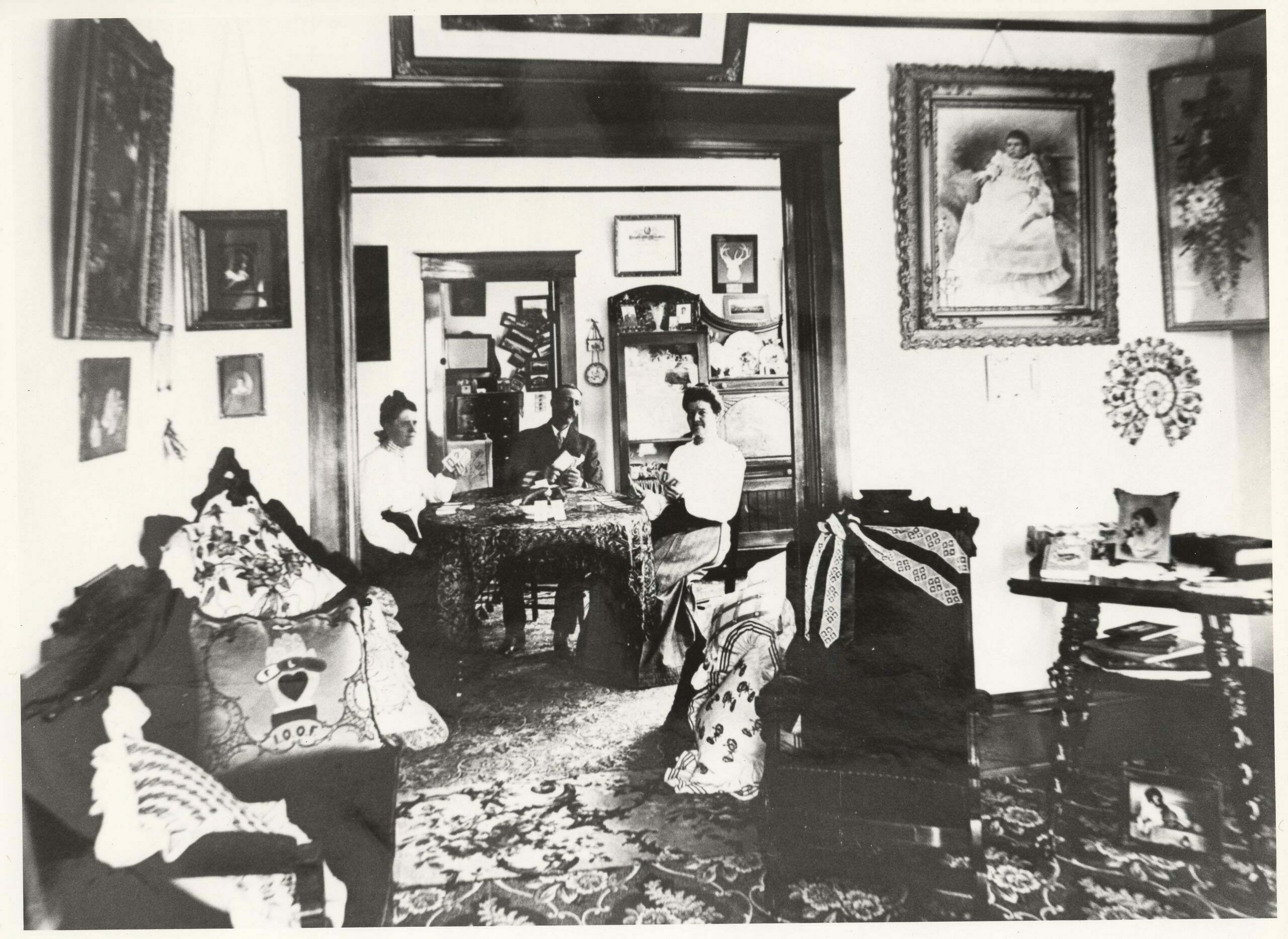 Late 19th century middle class living is evident in this Salida home, believed to be that of local photographer N. W. Meigs. The family plays cards amid busy decorations that include chair drapes, stacked pillows, heavy framed photographs and dark wood furnishings.  (1)