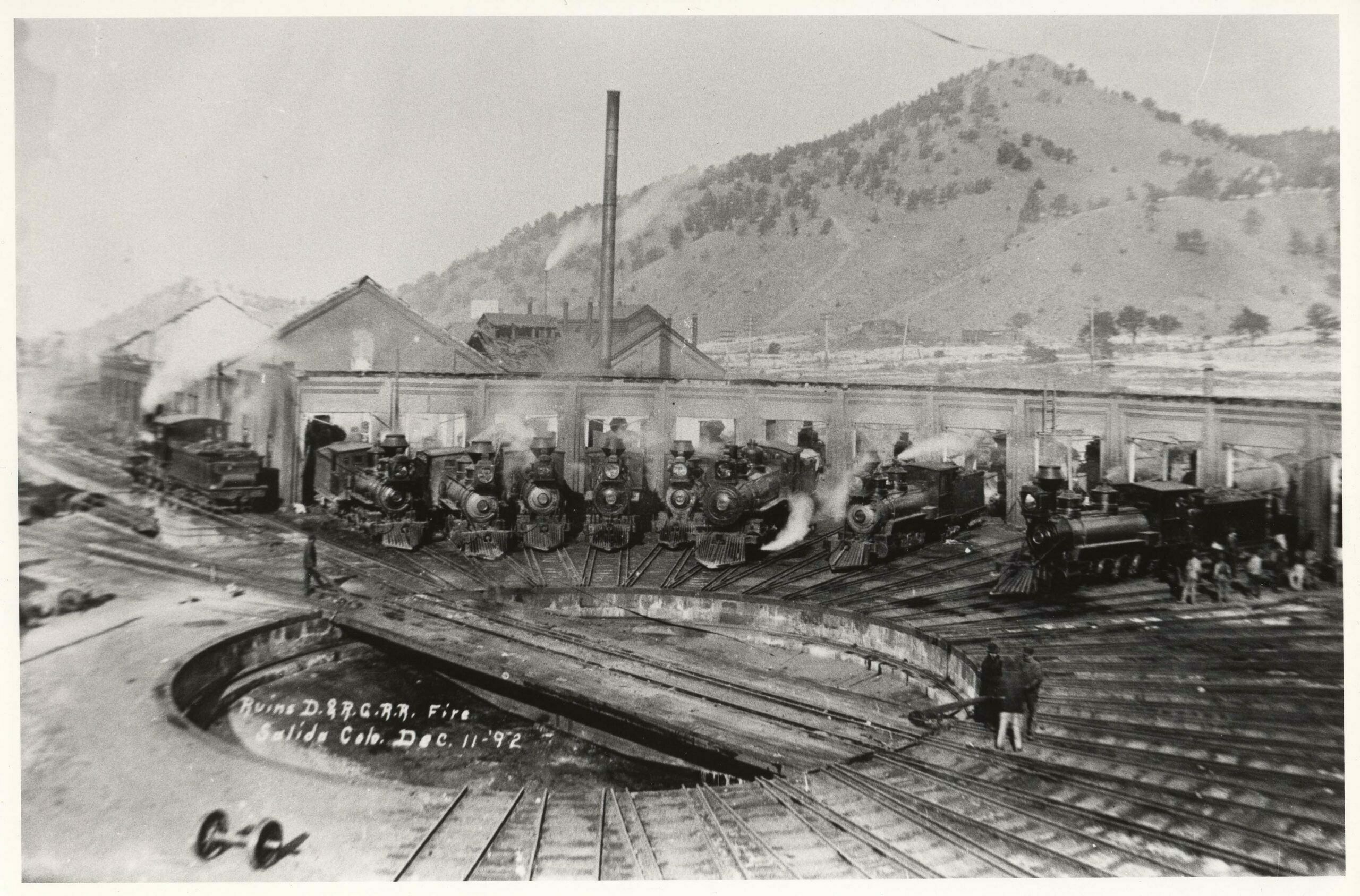 Shortly after the disastrous 1892 fire, locomotive servicing and repairs had to be done out in the open, in front of the roundhouse ruins. From this angle, the machine shop was at the left, and the charred boiler-house roof was visible beside the stack. Prior to the fire, the arch in the roundhouse doors had been bricked up. The 62-foot turntable was still in use. A standard-gauge switch engine, an 1890 Class 113 (C-28) Baldwin 2-8-0 in the 600-series, stood over the ashpit. Spotted on the roundhouse leads, from left to right, were Engines 227, 283, 400 (?), 401, 44, 530 (standard-gauge), 407 and 403. Burned hulks of locomotives remained in the remnants of the roundhouse.  (2)